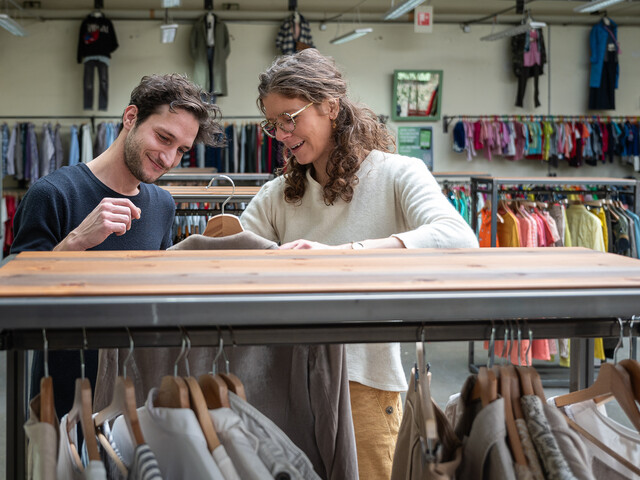 Zwei Personen stöbern fröhlich durch Kleidungsstücke in einem Second-Hand-Shop mit Kleiderständern und Mode im Hintergrund.