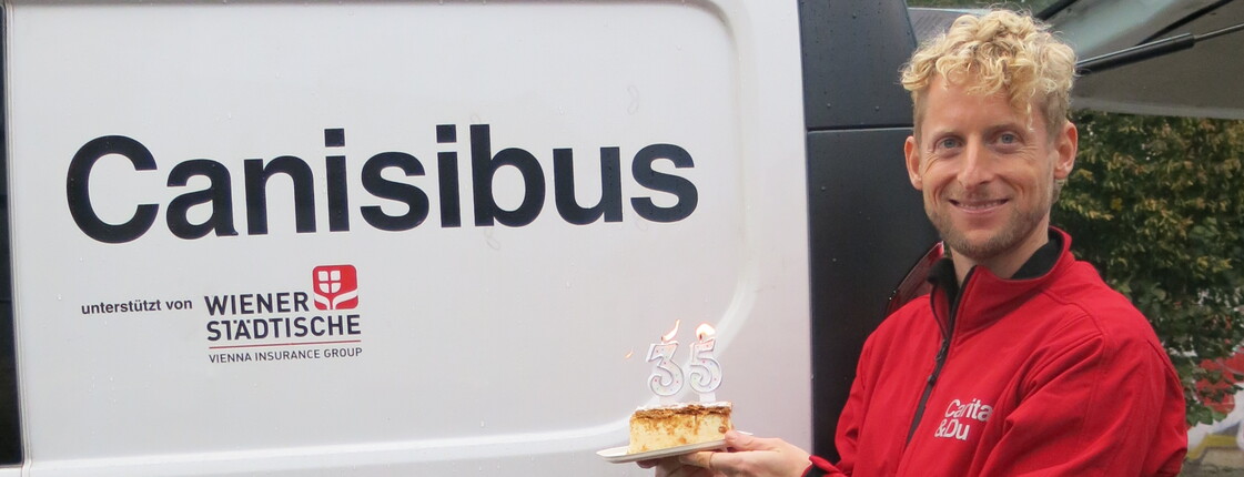 Mitarbeitender hält Kuchen mit Kerzen „35“ vor Canisibus-Fahrzeug der Caritas in Wien
